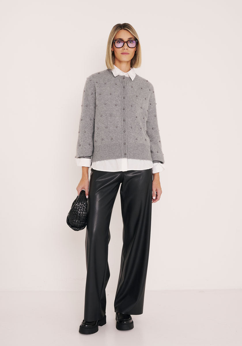 Eine Frau mit einem Bob-Haarschnitt und einer Brille trägt eine graue Strickjacke mit Perlenknöpfen von Princess Goes Hollywood über einem weißen Hemd, einer schwarzen, weit geschnittenen Lederhose und schwarzen Schuhen. In der Hand hält sie eine schwarze, strukturierte Clutch vor einem schlichten weißen Hintergrund.