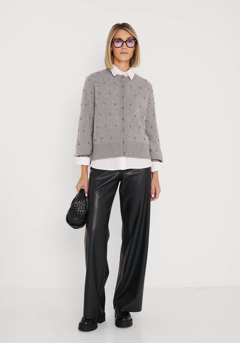 Eine Frau mit glattem blondem Haar und Brille steht vor einer weißen Wand. Sie trägt eine Princess Goes Hollywood Wollmix Strickjacke über einer weißen Bluse, eine schwarze Lederhose mit weitem Bein, schwarze Schuhe und eine kleine schwarze Handtasche mit Struktur.