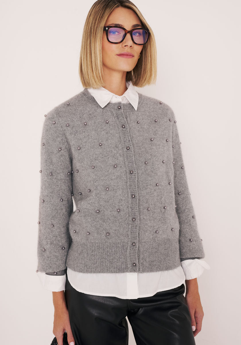 Eine selbstbewusste Frau mit glattem, schulterlangem blondem Haar und einer schwarz umrandeten Brille trägt ein weißes Hemd und eine graue Strickjacke mit Perlenknöpfen von Princess Goes Hollywood über einer schwarzen Lederhose, die vor einem hellen Hintergrund steht.