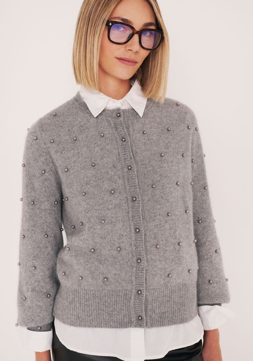 Eine Person mit glattem, schulterlangem blondem Haar und Brille trägt eine graue Princess Goes Hollywood Strickjacke mit kleinen perlenartigen Perlen über einem weißen Hemd mit Kragen und blickt mit sanftem Gesichtsausdruck in die Kamera vor einem schlichten hellen Hintergrund.