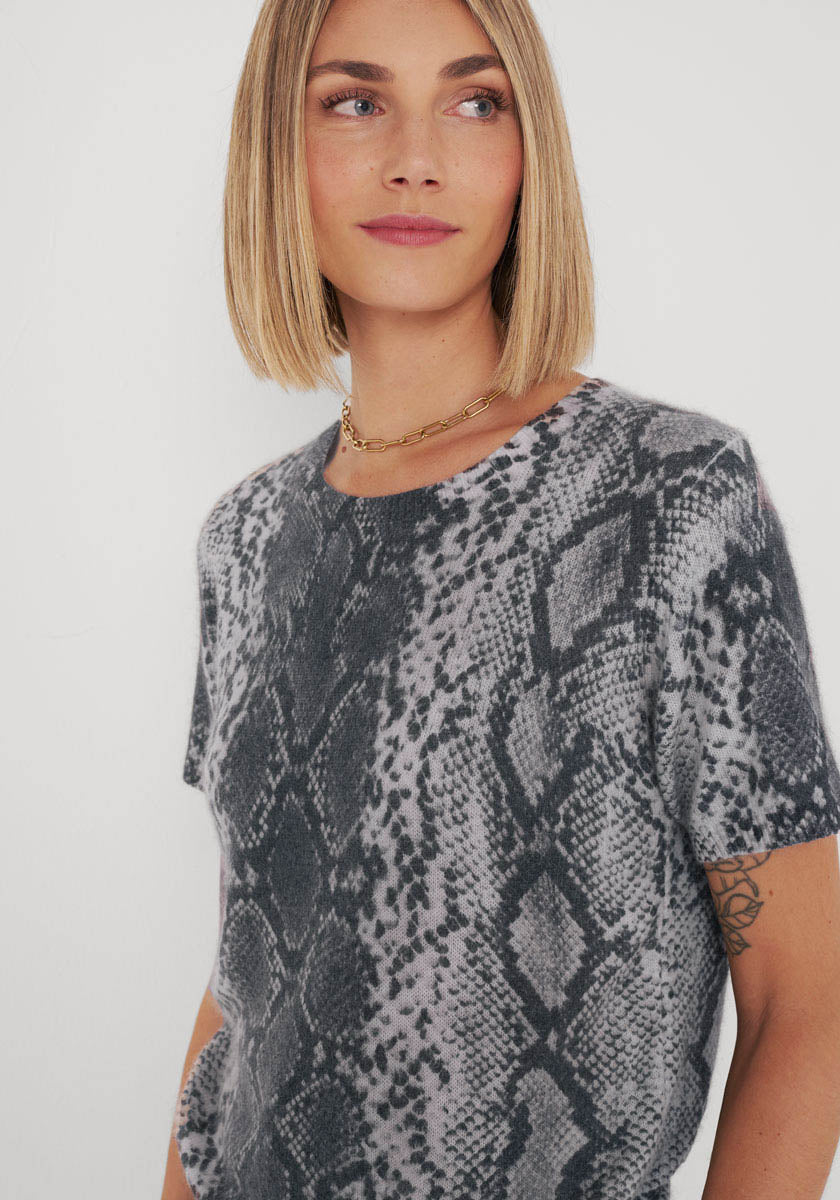 Eine Frau mit glattem, schulterlangem blondem Haar trägt ein kurzärmeliges Princess Goes Hollywood-Top mit Schlangenaufdruck und eine goldene Halskette. Sie hat ein leichtes Lächeln, ein neutrales Make-up, ein Oberarmtattoo und steht vor einem schlichten weißen Hintergrund.