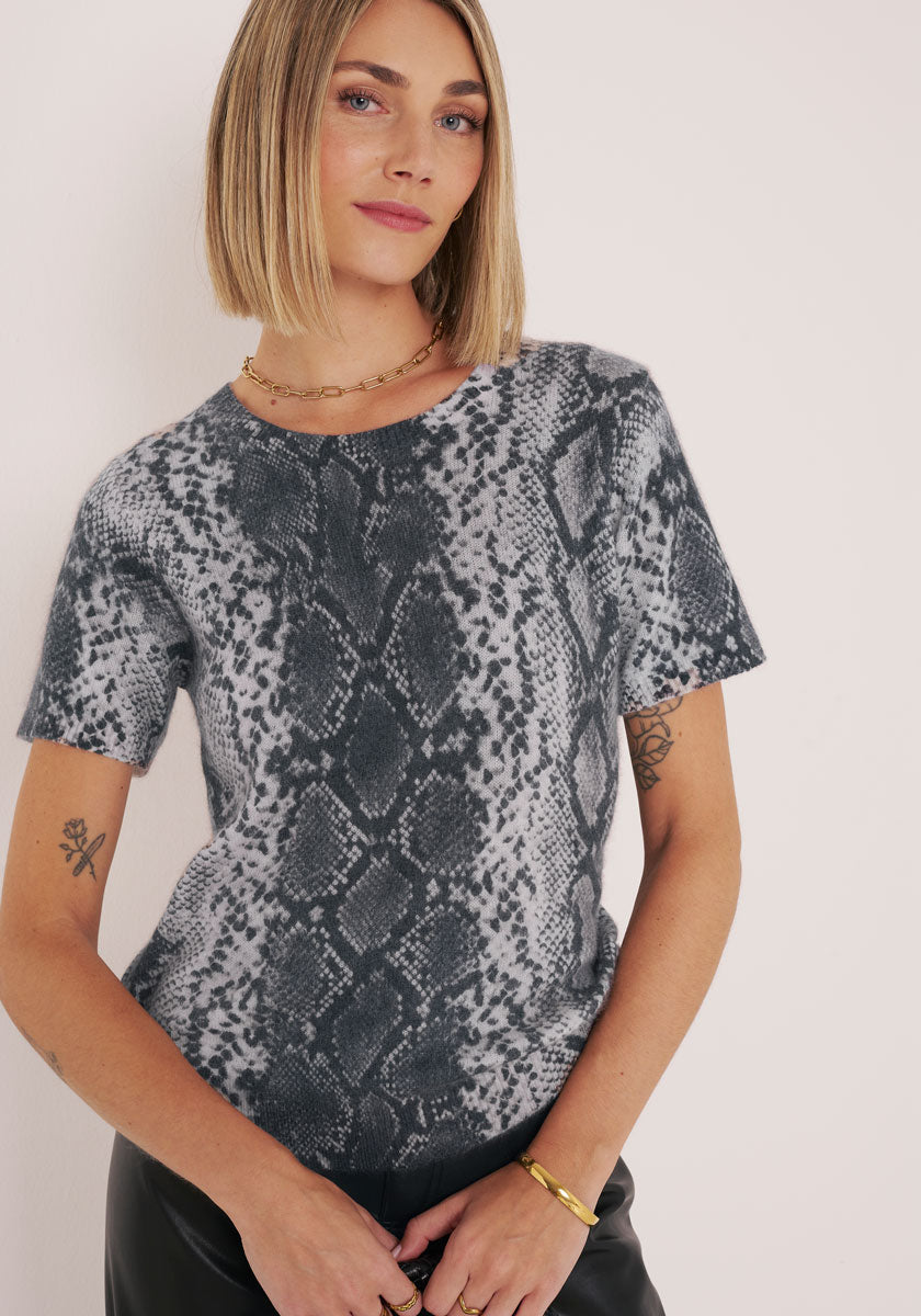 Eine Frau mit blondem Bob trägt ein gewagtes, modernes kurzärmeliges Oberteil mit Schlangendruck von Princess Goes Hollywood, gepaart mit kleinen Tattoos, einer goldenen Kettenhalskette und einem Armband. Sie steht selbstbewusst vor einem hellen Hintergrund.