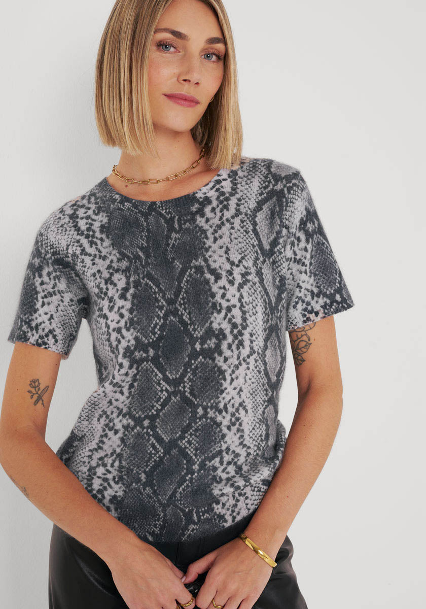 Eine Frau mit glattem, schulterlangem blondem Haar trägt einen grau-schwarzen Snake-Print Strickpullover von Princess Goes Hollywood. Sie hat helle Haut, Tattoos und Goldschmuck. Sie steht vor einem weißen Hintergrund und schaut selbstbewusst in die Kamera.