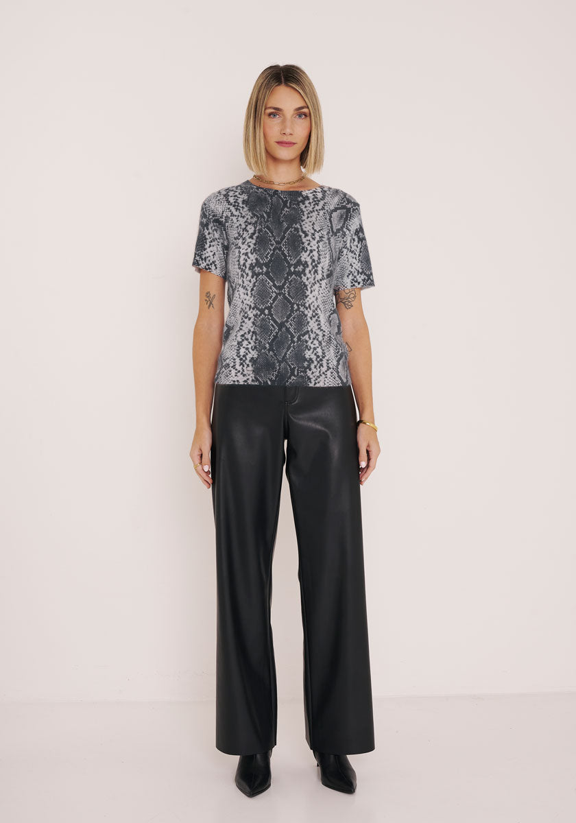 Eine Frau mit kinnlangem Bob steht vor einer weißen Wand. Sie trägt ein graues Oberteil mit Schlangenaufdruck von Princess Goes Hollywood und eine schwarze Hose aus Kunstleder mit weitem Bein, dazu spitze schwarze Schuhe. Ihre Arme ruhen entspannt an ihren Seiten.