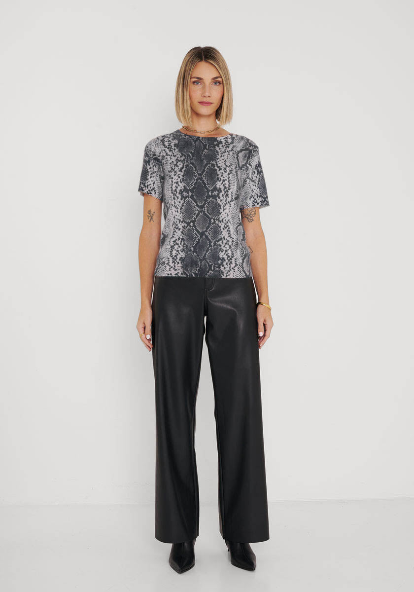 Eine Frau mit blondem Bob steht vor einem weißen Hintergrund und trägt ein graues T-Shirt mit Schlangenaufdruck von Princess Goes Hollywood, eine schwarze, weit geschnittene Lederhose und schwarze, spitze Schuhe. Auf ihrem linken Unterarm sind Tätowierungen zu sehen, während sie neutral in die Kamera blickt.