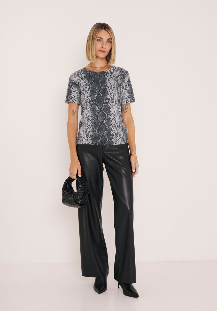 Eine Frau mit glattem blondem Haar und neutralem Gesichtsausdruck steht vor einer weißen Wand. Sie trägt ein graues kurzärmeliges Oberteil mit Schlangendruck von Princess Goes Hollywood, eine schwarze Lederhose mit weitem Bein, spitze schwarze Stiefel und eine gewebte schwarze Handtasche.