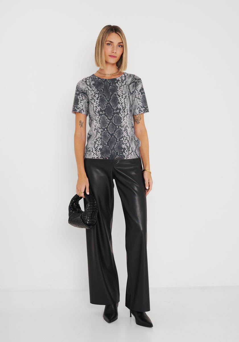 Eine Frau mit blondem Bob steht vor einer weißen Wand. Sie trägt ein graues kurzärmeliges Oberteil mit Schlangenaufdruck von Princess Goes Hollywood, eine schwarze Lederhose und schwarze spitze Stiefel. Sie hält eine kleine schwarze Handtasche in ihrer linken Hand und zeigt eine Tätowierung auf ihrem rechten Unterarm.