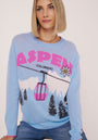 Eine Frau mit blondem Haarschopf trägt einen hellblauen Princess Goes Hollywood Strickpullover mit fettgedrucktem rosa "ASPEN", schwarzem "COLORADO", schneebedeckten Bergen, einer rosafarbenen Gondel, Kiefern und einer Edelweißblüte, die vor einem unifarbenen Hintergrund sitzt.
