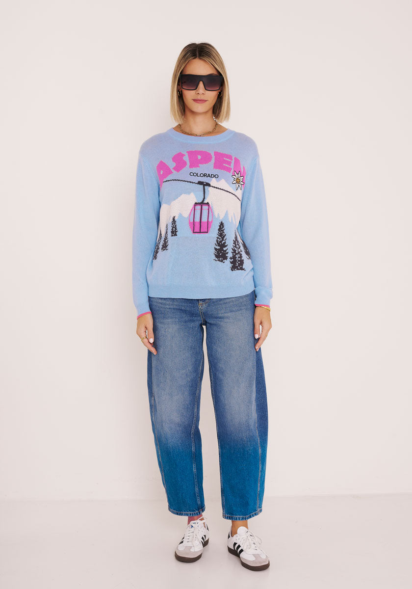 Eine Frau steht vor einem weißen Hintergrund und trägt eine dunkle Sonnenbrille, blaue Jeans, weiße Turnschuhe mit schwarzen Akzenten und einen hellblauen Aspen Colorado Strickpullover mit Berg-, Seilbahn- und Baumdesign von Princess Goes Hollywood.