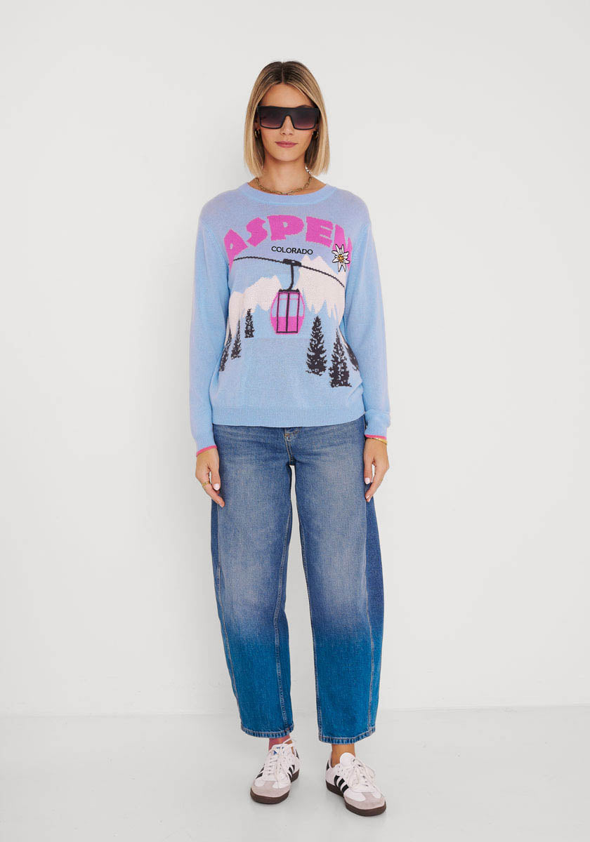 Eine Frau steht vor einer weißen Wand und trägt eine Sonnenbrille, eine blaue, locker sitzende Jeans, weiße Turnschuhe mit dunklen Akzenten und einen hellblauen Princess Goes Hollywood Aspen Colorado Strickpullover mit einer Berg-, Baum- und Gondelgrafik.