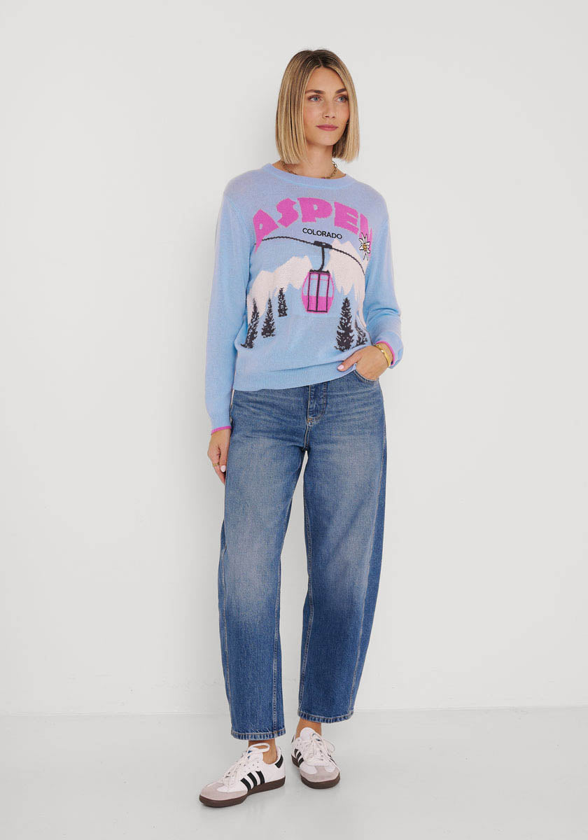 Eine Frau mit glattem, schulterlangem blondem Haar steht vor einer weißen Wand und trägt einen hellblauen Princess Goes Hollywood Aspen Colorado Strickpullover mit Berg- und Gondelgrafik, blaue Jeans mit Bündchen und klassische weiße Adidas-Sneaker mit schwarzen Streifen.