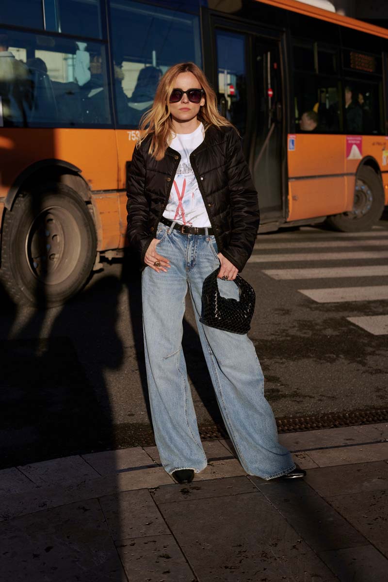 Eine Frau steht auf einer Straße in der Nähe eines orangefarbenen Busses. Sie trägt eine übergroße Sonnenbrille, eine schwarze Pufferjacke, ein Princess Goes Hollywood T-Shirt mit Skidruck aus Baumwolle, hellblaue Jeans mit weitem Bein, schwarze Schuhe und eine schwarze strukturierte Handtasche.