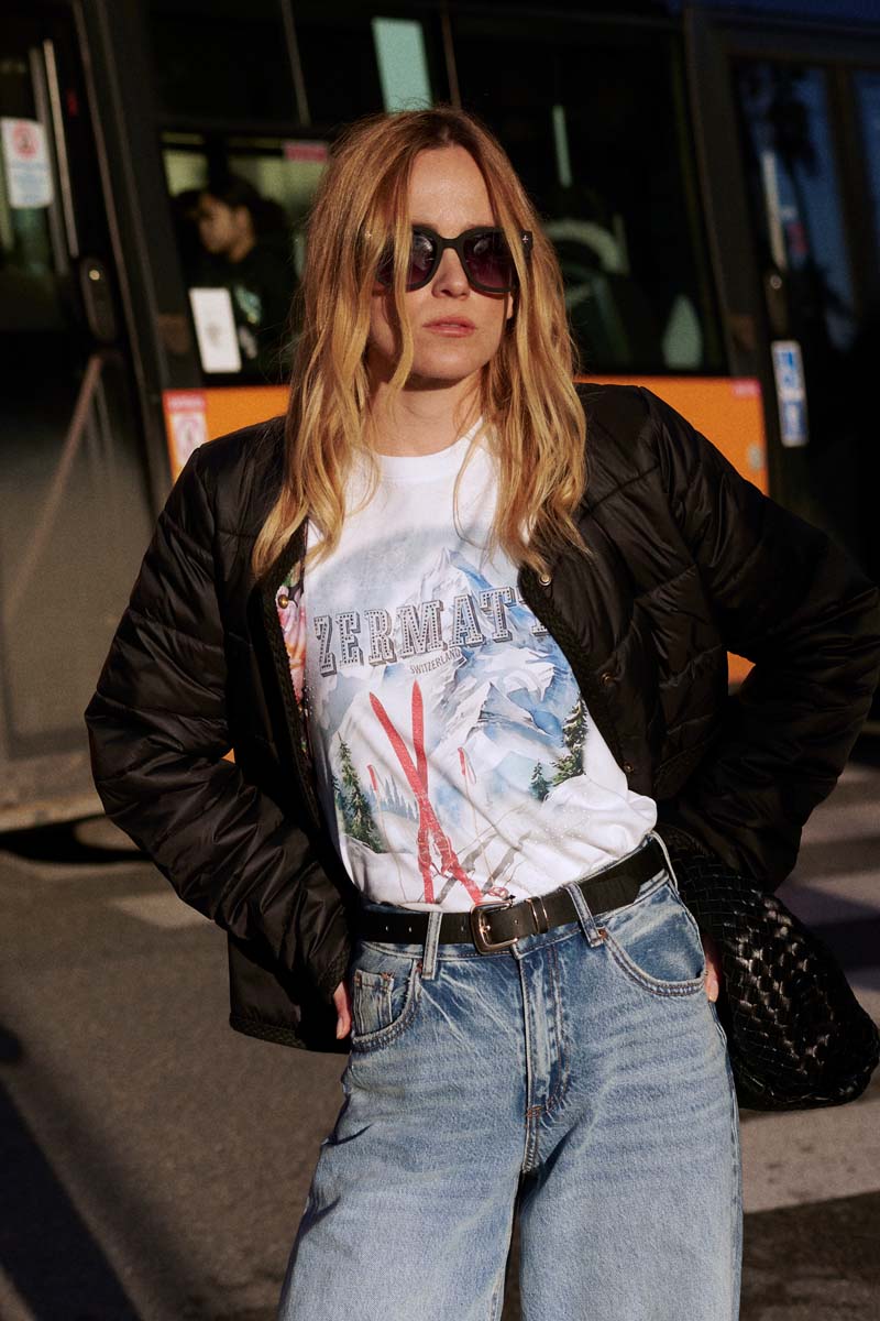 Eine Frau mit langen blonden Haaren, die eine schwarze Sonnenbrille, ein weißes Princess Goes Hollywood Ski-Print T-Shirt Baumwolle, eine schwarze Steppjacke und Blue Jeans trägt, steht im Freien im Sonnenlicht. Hinter ihr ist ein Stadtbus geparkt, in dem eine Person zu sehen ist.