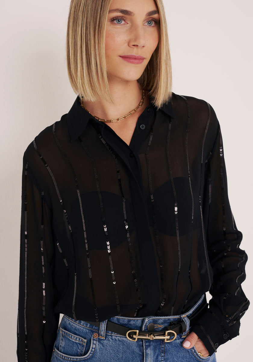 Eine Frau mit glattem blondem Haar trägt eine transparente Hemdbluse mit Streifen mit Pailletten von Princess Goes Hollywood über einem schwarzen BH, gestylt mit blauen Jeans, einem schwarzen Gürtel mit goldener Schnalle und einer goldenen Kettenhalskette. Sie dreht sich leicht und lächelt sanft.