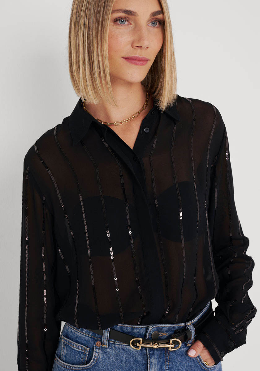 Eine Frau mit glattem blondem Haar trägt eine transparente Hemdbluse mit Paillettenbesatz und Streifen von Princess Goes Hollywood über einem schwarzen BH, kombiniert mit Blue Jeans, schwarzem Gürtel mit goldener Schnalle und goldener Kette vor hellem Hintergrund.