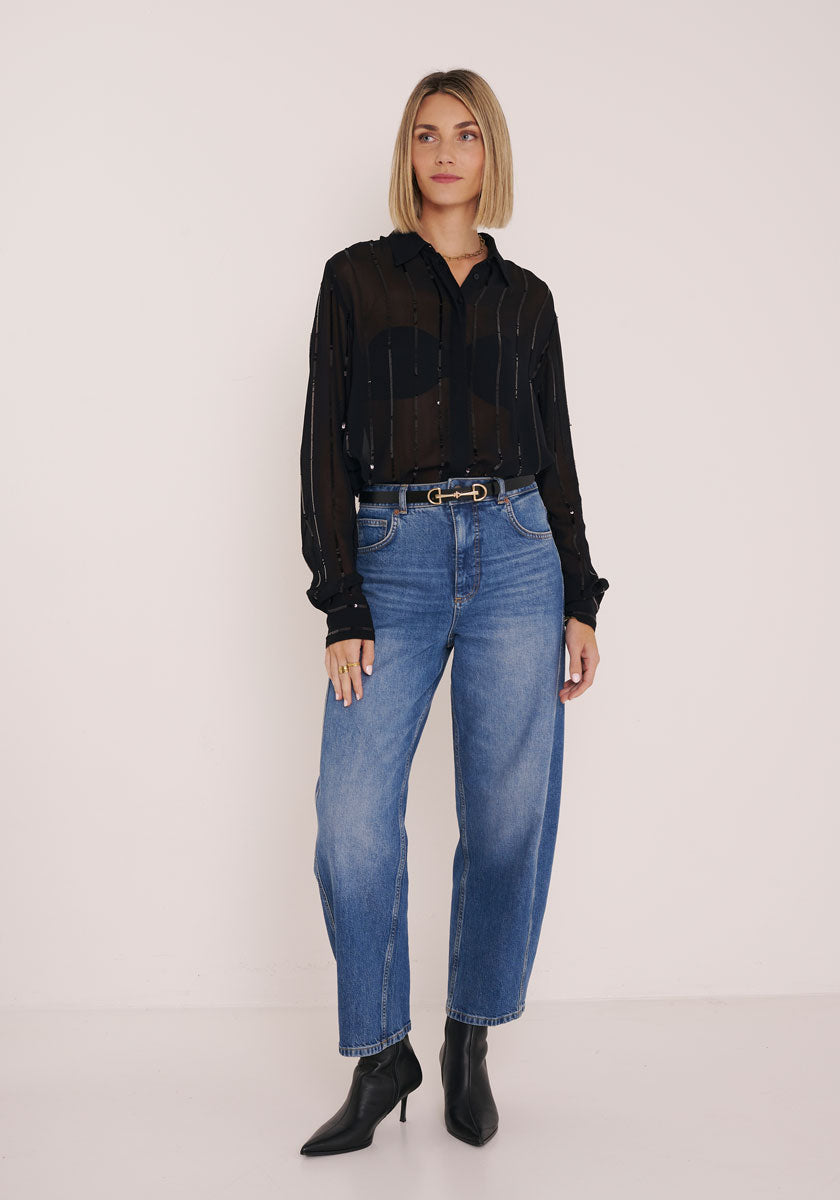 Eine Frau mit blondem Bob steht vor einer schlichten Wand und trägt eine transparente Hemdbluse von Princess Goes Hollywood über einem schwarzen BH, blaue High-Waisted-Jeans, einen schwarzen Gürtel mit goldener Schnalle und schwarze spitze Ankle Boots für einen schicken Look.