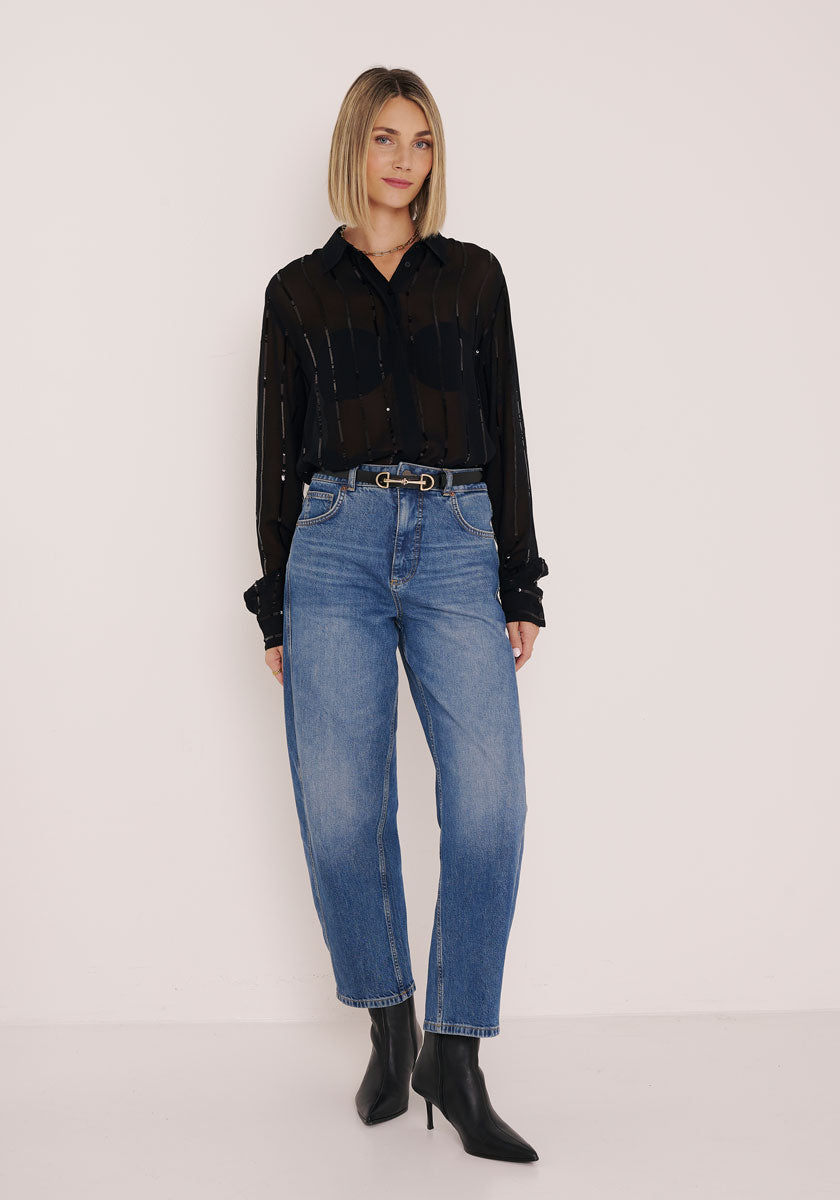 Eine Frau mit geradem, schulterlangem blondem Bob trägt eine transparente Princess Goes Hollywood Hemdbluse über einem schwarzen BH, die in eine hoch taillierte Bluejeans mit schwarzem Gürtel und spitzen schwarzen Stiefeletten gesteckt ist, und steht vor einer schlichten hellen Wand.