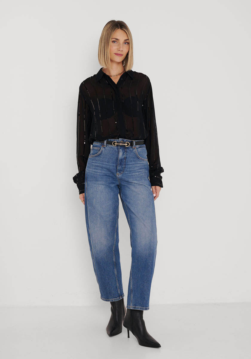 Eine Frau mit glattem blondem Haar und neutralem Gesichtsausdruck steht vor einer weißen Wand und trägt eine schwarze transparente Hemdbluse von Princess Goes Hollywood, blaue High-Waisted-Jeans, einen schwarzen Gürtel mit goldener Schnalle und schwarze hochhackige Stiefeletten.