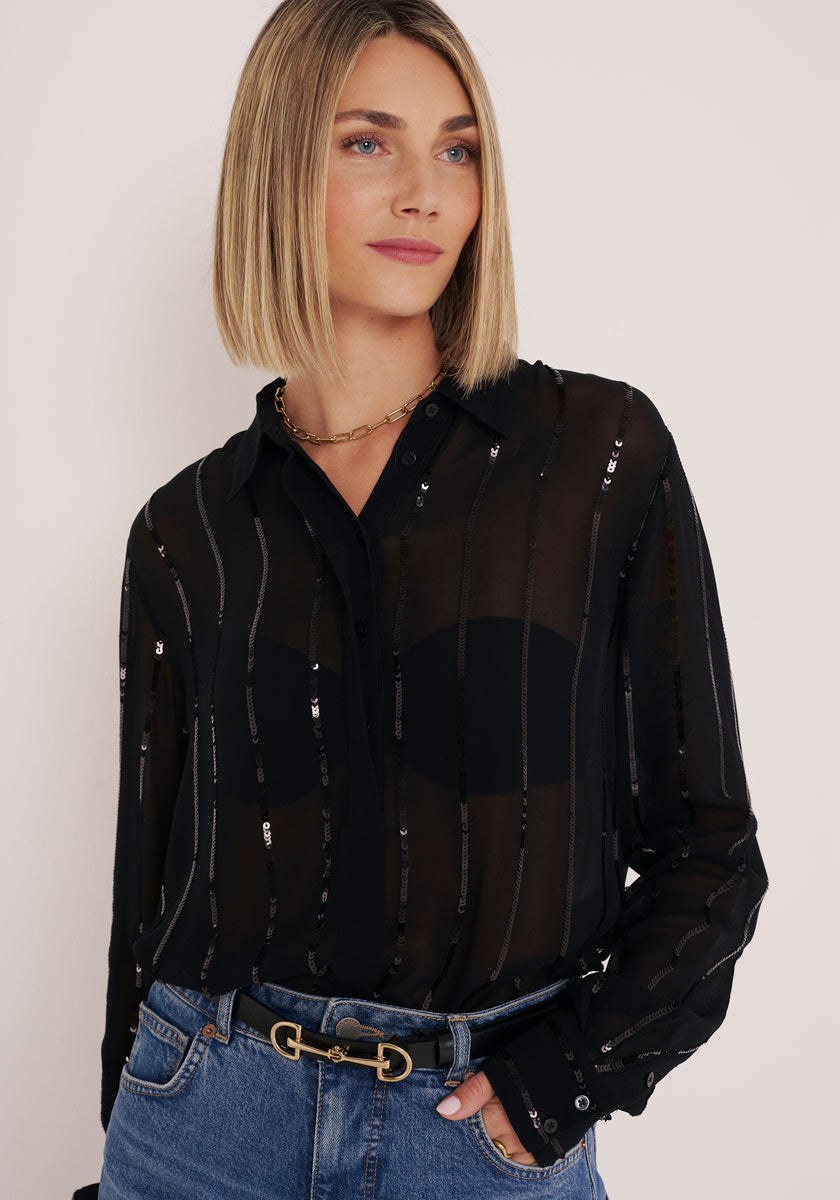 Eine Frau mit blondem Bob trägt eine transparente Hemdbluse mit Streifen und Pailletten von Princess Goes Hollywood über einem schwarzen BH, dazu blaue Jeans und einen schwarzen Gürtel mit goldener Schnalle. Sie steht vor hellem Hintergrund.