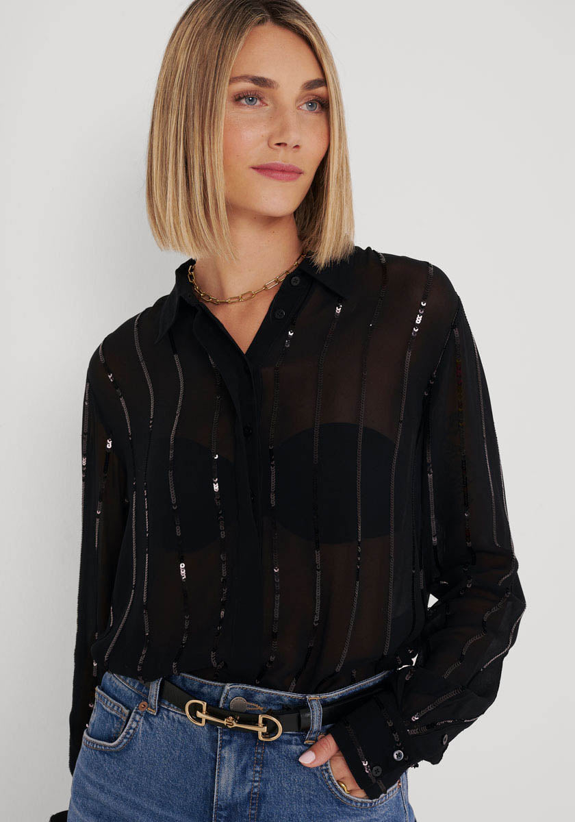 Eine Frau mit glattem, schulterlangem blondem Haar trägt eine transparente, gestreifte Bluse mit Paillettenbesatz von Princess Goes Hollywood über einem schwarzen BH, dazu Blue Jeans, einen schwarzen Gürtel und eine goldene Halskette. Sie steht vor einem hellen Hintergrund und blickt zur Seite.