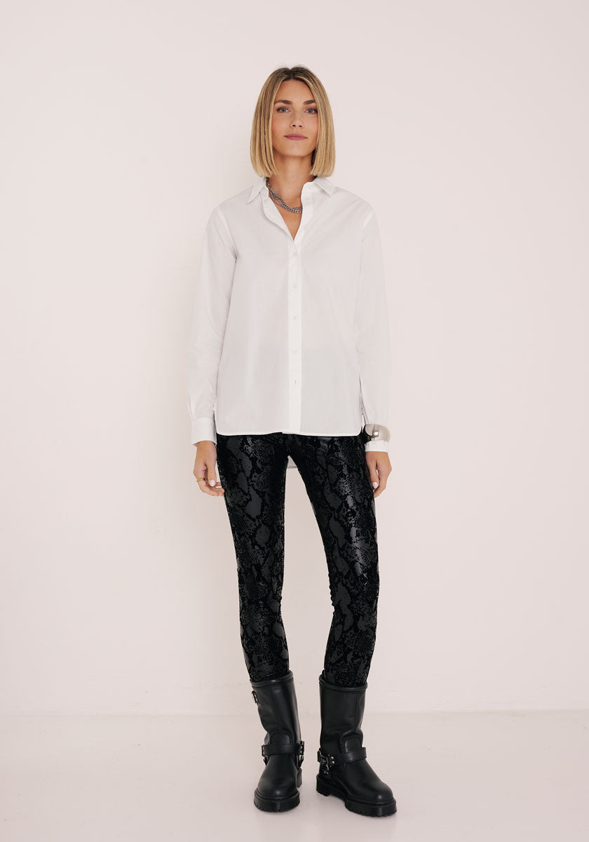 Eine Frau mit blondem Bob steht vor einer weißen Wand und trägt ein weißes Princess Goes Hollywood-Knopfhemd, schwarze Kunstleder-Leggings mit Schlangendruck und elastischem Bund sowie schwarze, klobige Stiefel. Sie hat einen neutralen Gesichtsausdruck und entspannte Arme.