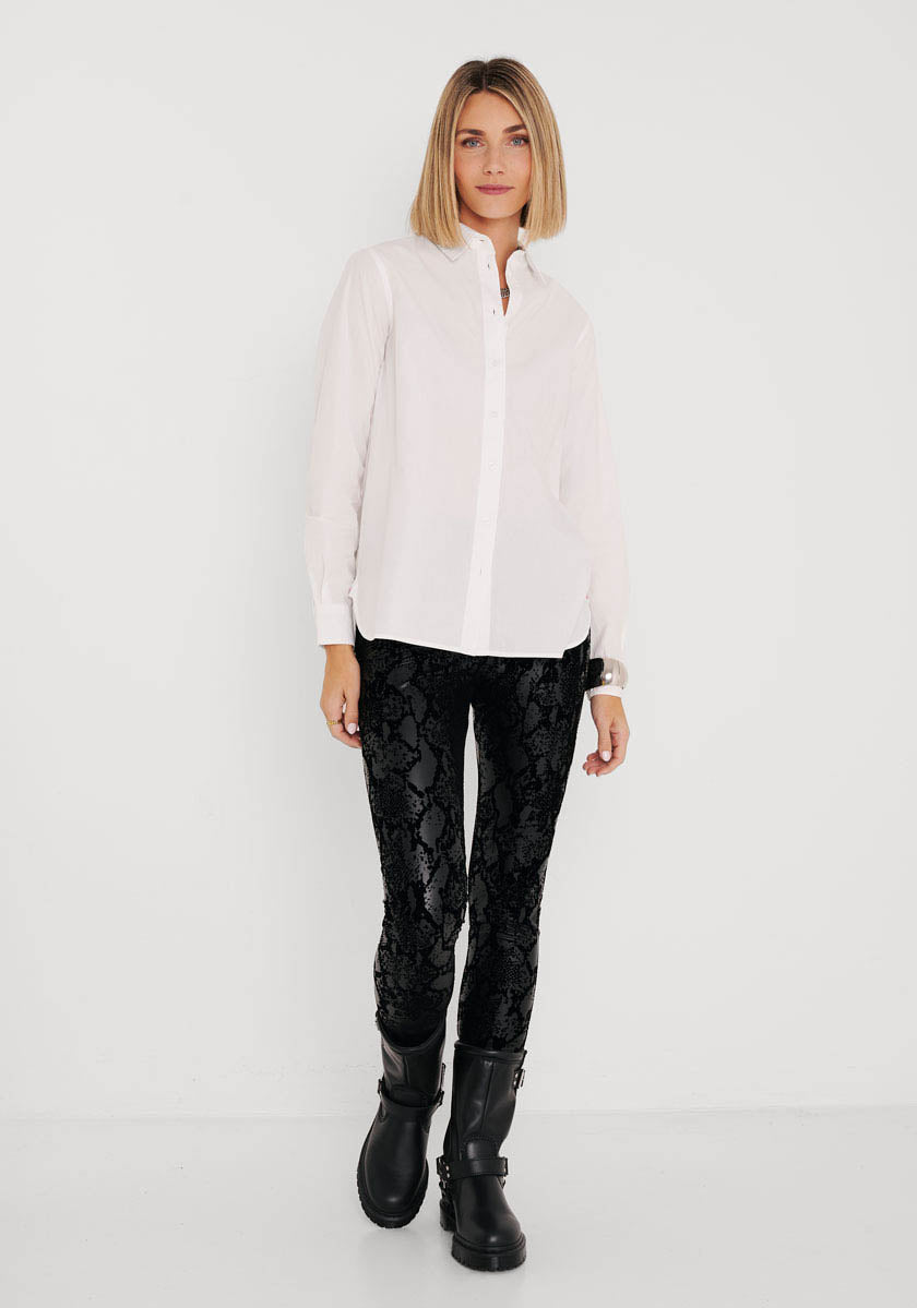 Eine Frau mit glattem blondem Haar trägt ein weißes Button-up-Hemd, Princess Goes Hollywood, schwarze Kunstlederleggings mit Schlangenaufdruck und elastischem Bund sowie schwarze Stiefel. Sie steht mit entspanntem Gesichtsausdruck vor einem schlichten weißen Hintergrund.