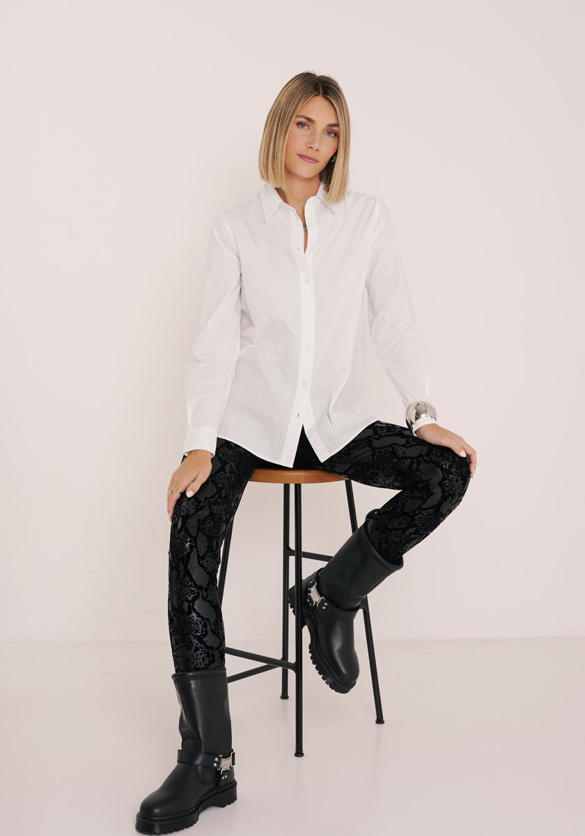 Eine Frau mit Bobfrisur sitzt auf einem hohen Hocker vor einem schlichten weißen Hintergrund. Sie trägt eine Princess Goes Hollywood weiße Hemdbluse aus Baumwolle, eine gemusterte schwarze Samthose und schwarze klobige Stiefel. Sie wirkt ruhig und selbstbewusst, einen Arm auf ihr Knie gestützt.