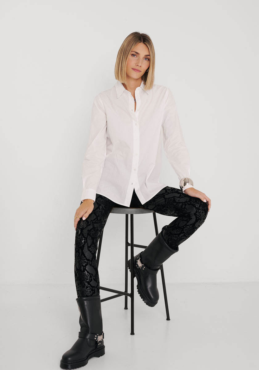 Eine Frau mit blondem Bubikopf sitzt auf einem schwarzen Hocker vor einem weißen Hintergrund. Sie trägt ein weißes, geknöpftes Princess Goes Hollywood-Hemd, Kunstleder-Leggings mit Schlangenaufdruck und elastischer Taille sowie klobige schwarze Stiefel und schaut ruhig in die Kamera.