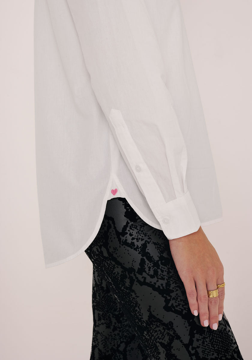 Eine Person trägt eine lockere weiße Baumwoll-Hemdbluse von Princess Goes Hollywood mit kleinem pinkem Herzdetail am Saum zu schwarzen strukturierten Hosen. Mehrere goldene Ringe schmücken ihre Hand. Der Hintergrund ist schlicht und hell.