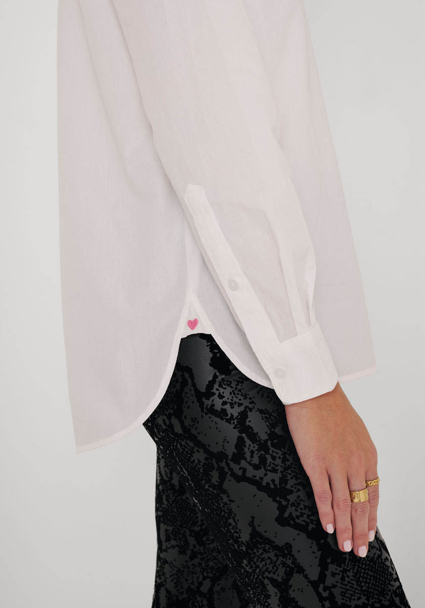 Eine Person trägt eine lockere, weiße Hemdbluse aus Baumwolle mit Herzdetails von Princess Goes Hollywood, hochgekrempelten Ärmeln, dazu einen schwarz-grauen Rock und goldene Ringe. Ihre Hand ruht entspannt an der Seite.