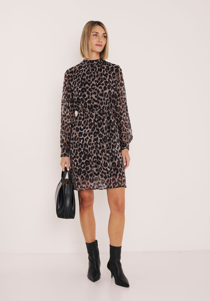 Eine Frau steht in einem knielangen, langärmeligen Princess Goes Hollywood Wild-Leo-Print-Kleid vor einer weißen Wand, hält eine schwarze Handtasche in der Hand und trägt schwarze Ankle Boots. Ihr glattes, kinnlanges Haar unterstreicht ihren selbstbewussten Stil.