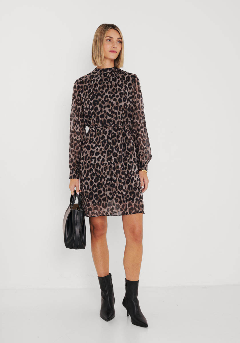 Eine Frau posiert vor einer weißen Wand in einem knielangen, langärmeligen Princess Goes Hollywood Wild-Leo-Print-Kleid. Dazu trägt sie eine schwarze Handtasche und schwarze Stiefeletten. Ihr glattes, schulterlanges blondes Haar rundet das schicke Outfit ab.