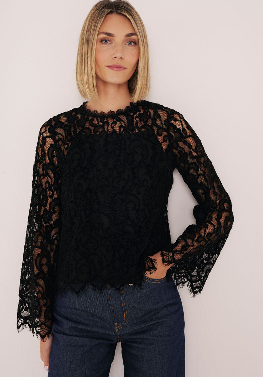 Eine Frau mit geradem, blondem Bob trägt eine figurbetonte Bluse aus schwarzer Lochspitze mit Stehkragen von Princess Goes Hollywood über einem schwarzen Unterhemd und kombiniert sie mit einer dunkelblauen High-Waist-Jeans.