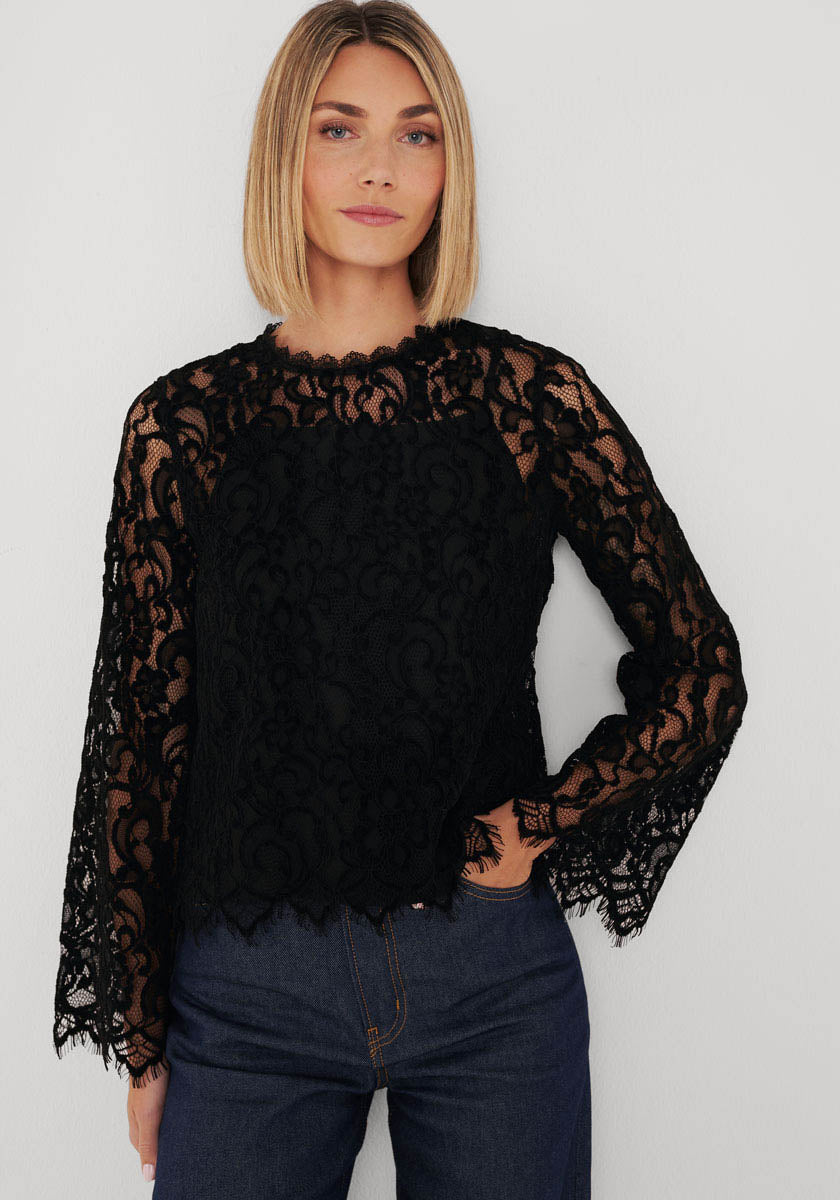 Eine Frau mit glattem, schulterlangem blondem Haar modelliert eine figurbetonte Bluse aus schwarzer Lochspitze mit Stehkragen von Princess Goes Hollywood, hoch taillierte blaue Jeans und ein schwarzes Unterteil vor weißem Hintergrund.