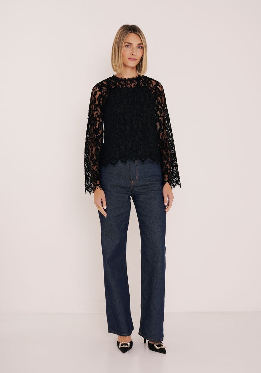 Eine Frau mit glattem, schulterlangem blondem Haar und neutralem Gesichtsausdruck steht vor einem weißen Hintergrund und trägt eine figurbetonte schwarze Ösenbluse mit Stehkragen von Princess Goes Hollywood, dunkelblaue Jeans mit geradem Bein und schwarze spitze Absätze.