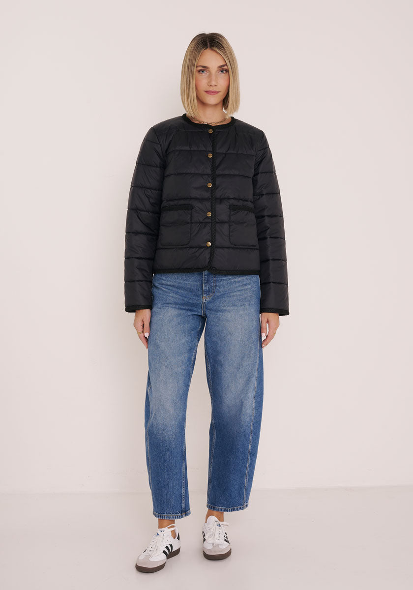 Eine Frau mit blondem Bob trägt eine schwarze gesteppte Kurzjacke mit Rundhalsausschnitt von Princess Goes Hollywood, dazu weite blaue Jeans und weiße Sneakers mit schwarzen Streifen. Sie steht vor einer hellen Wand und wirkt entspannt.