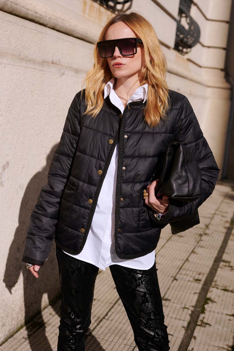 Eine Frau mit blondem Haar und einer großen Sonnenbrille trägt eine kurze Steppjacke mit goldenen Knöpfen, ein weißes Hemd und eine schwarze strukturierte Hose. Sie steht an einer cremefarbenen Wand im Sonnenlicht und im Schatten, die die städtische Umgebung hervorheben.