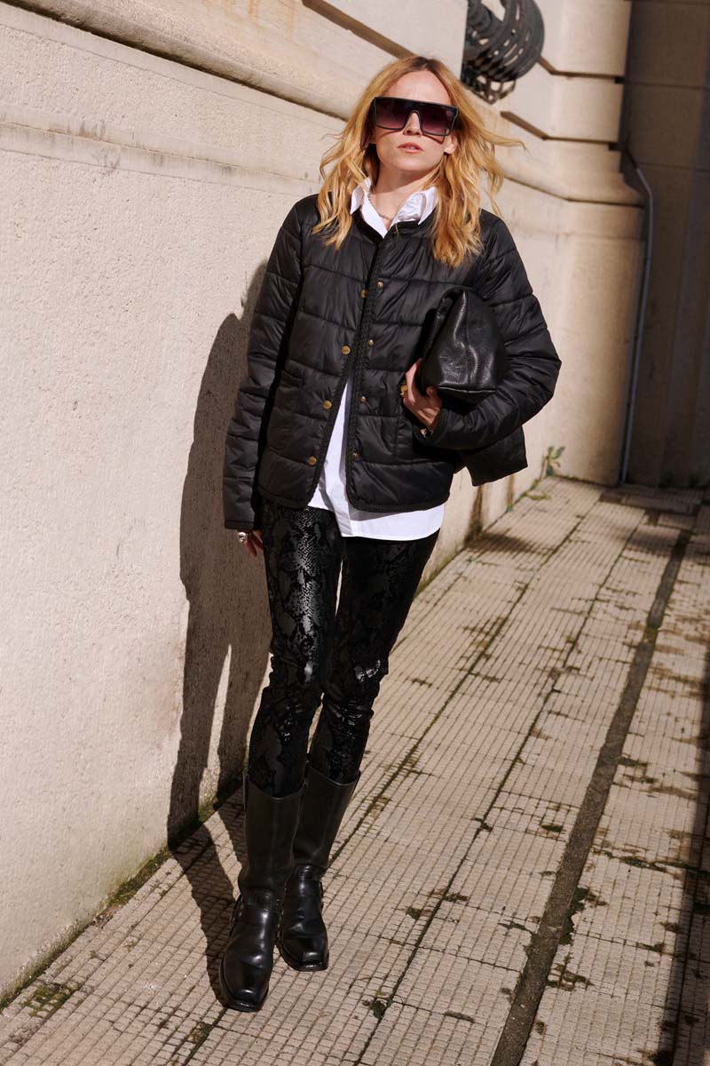 Eine blonde Frau mit Sonnenbrille trägt eine Princess Goes Hollywood gesteppte Kurzjacke mit Rundhalsausschnitt und goldfarbenen Druckknöpfen über einem weißen Hemd, schwarzen Spitzenleggins und schwarzen Stiefeln und hält eine große schwarze Clutch an einer beigen Steinmauer.