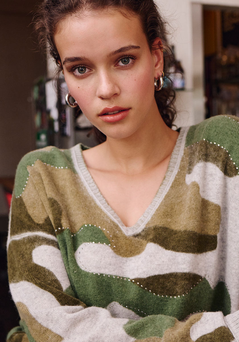 A young woman with curly brown hair wears hoop earrings and a Frogbox by Princess Camouflage-Print Merino Strickpullover with subtle rhinestone details. She has light makeup and looks directly at the camera, standing indoors with a neutral expression.