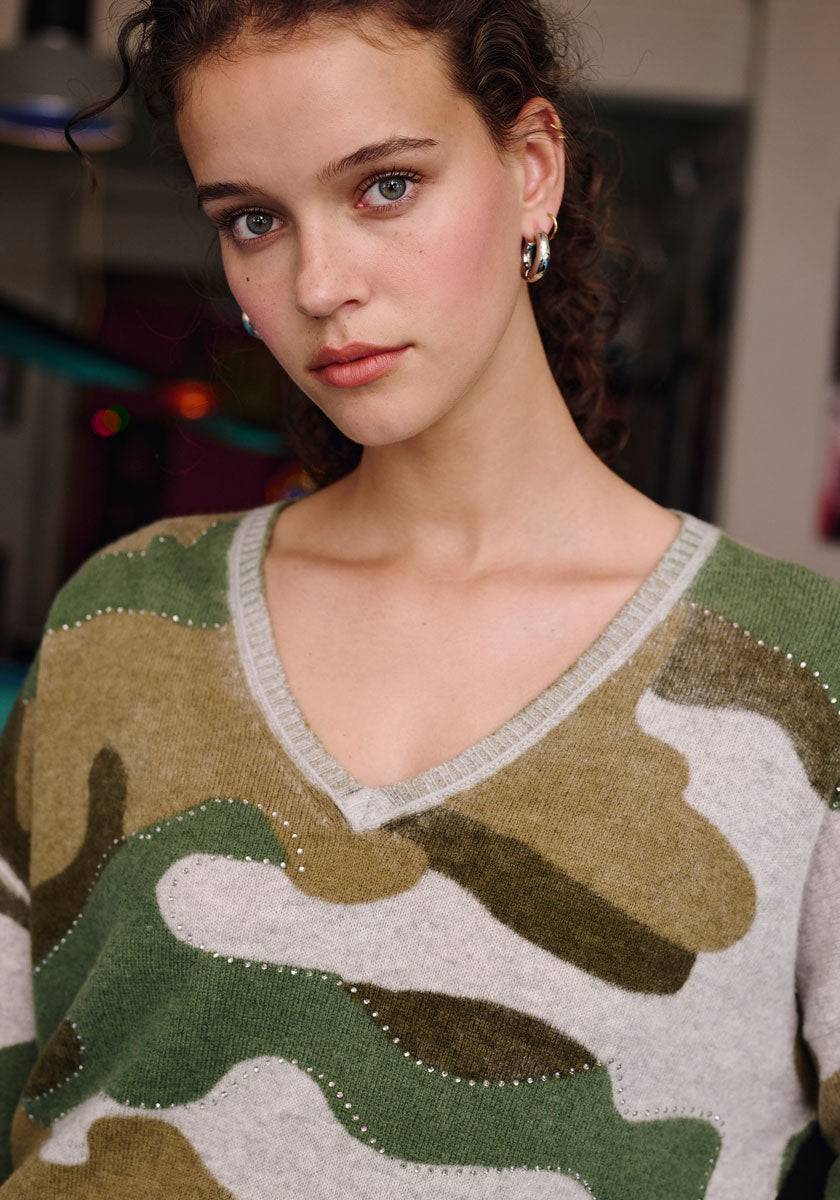 A young woman with light skin and curly brown hair wears a green and tan Camouflage-Print Merino Strickpullover by Frogbox by Princess, featuring small Strassverzierung. She accessorizes with silver hoop earrings and subtle makeup, gazing at the camera.