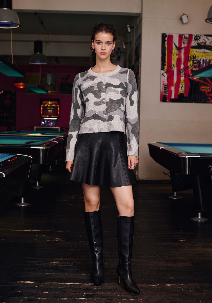 A woman stands indoors on a dark wooden floor between pool tables, wearing a gray camouflage Strickjacke by Frogbox by Princess, a black leather skirt, and black knee-high boots. Her hair is tied back; colorful art decorates the stylish setting.