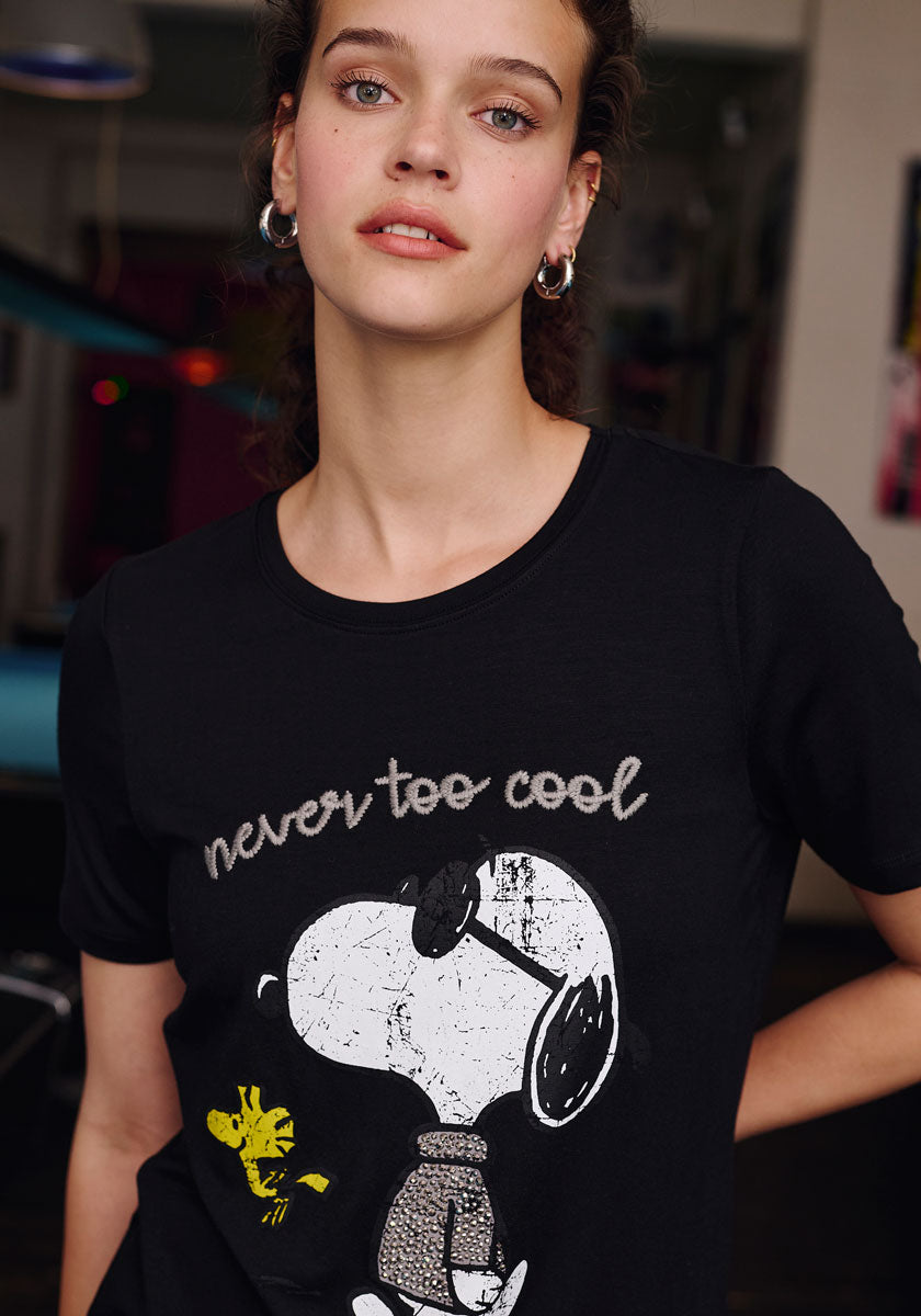 A young woman with light skin and curly brown hair wears hoop earrings and a black Frogbox by Princess Snoopy T-shirt with a crew neck. The shirt features Snoopy in sunglasses, Woodstock, and the text never too cool as she poses indoors.