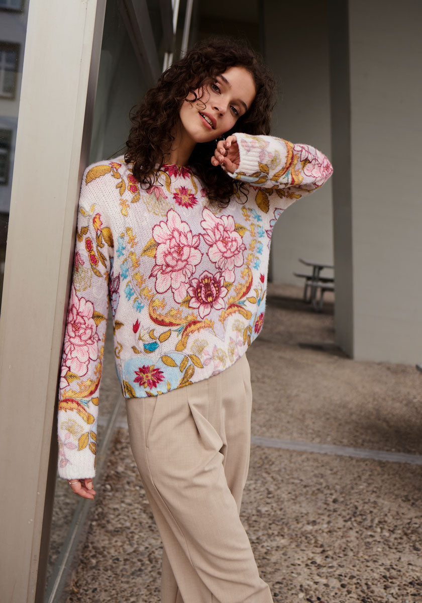 A woman with curly brown hair smiles softly as she leans against a wall outdoors, wearing a Princess Goes Hollywood Strickpullover with Merinoanteil in retro-blumen-print and high-waisted beige pants. A window and bench appear in the background.
