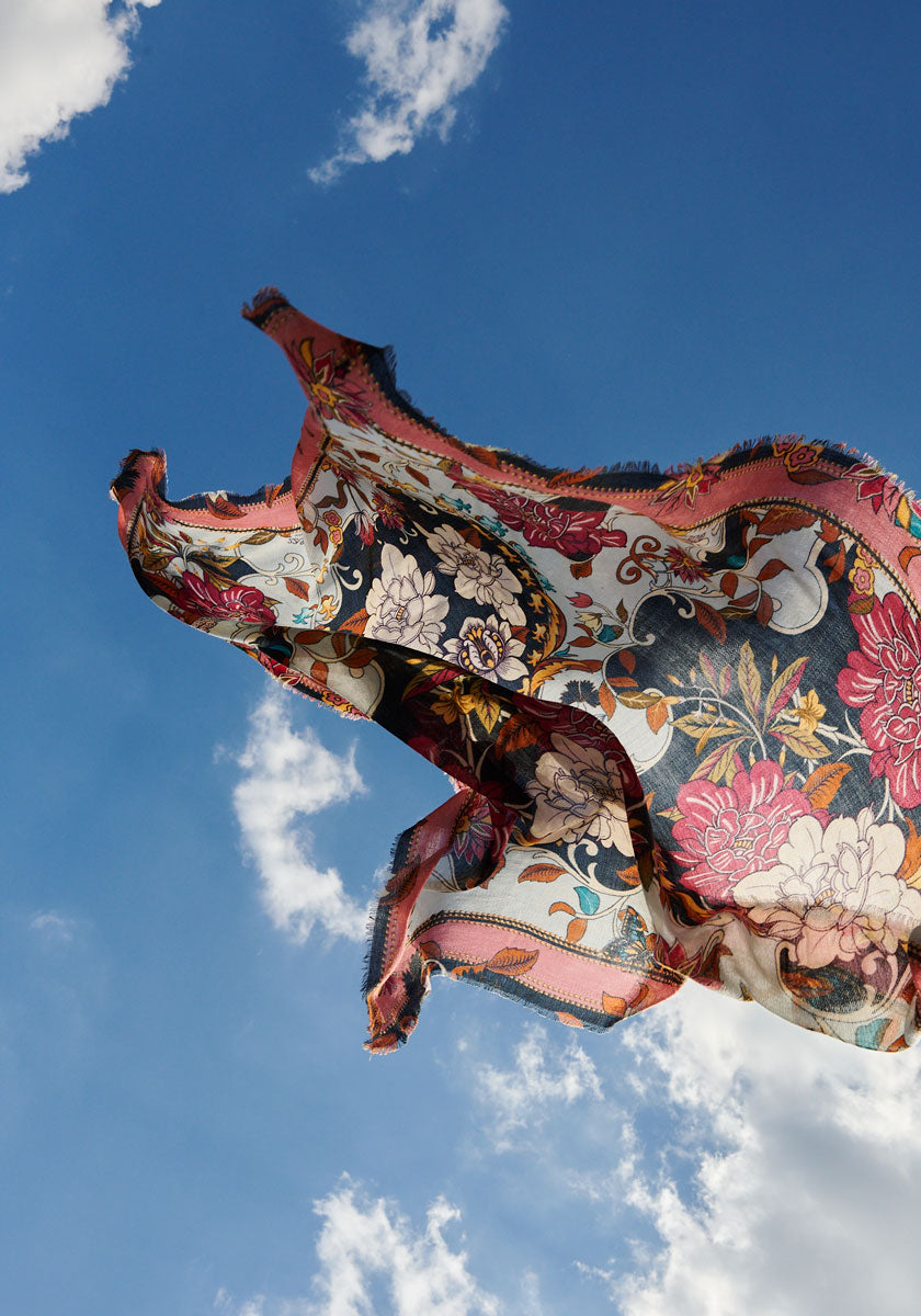 A colorful Princess Goes Hollywood Retro-Blumen-Print quadratisches Tuch with frayed edges billows gracefully against a bright blue sky dotted with white clouds, creating a dynamic sense of movement and contrast.
