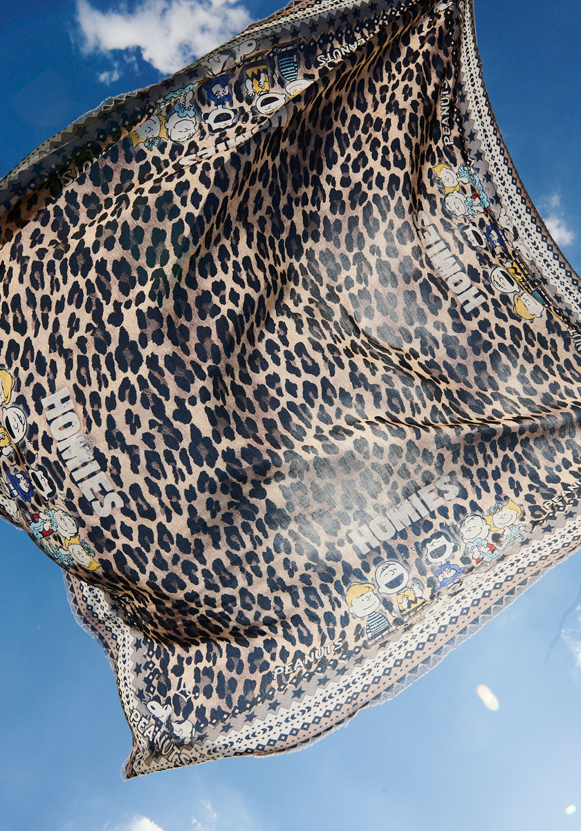 A Princess Goes Hollywood Glam-Leo-Print scarf featuring cartoon faces and HOMIES along the border billows against a bright blue sky with scattered clouds, seen from a low-angle view.