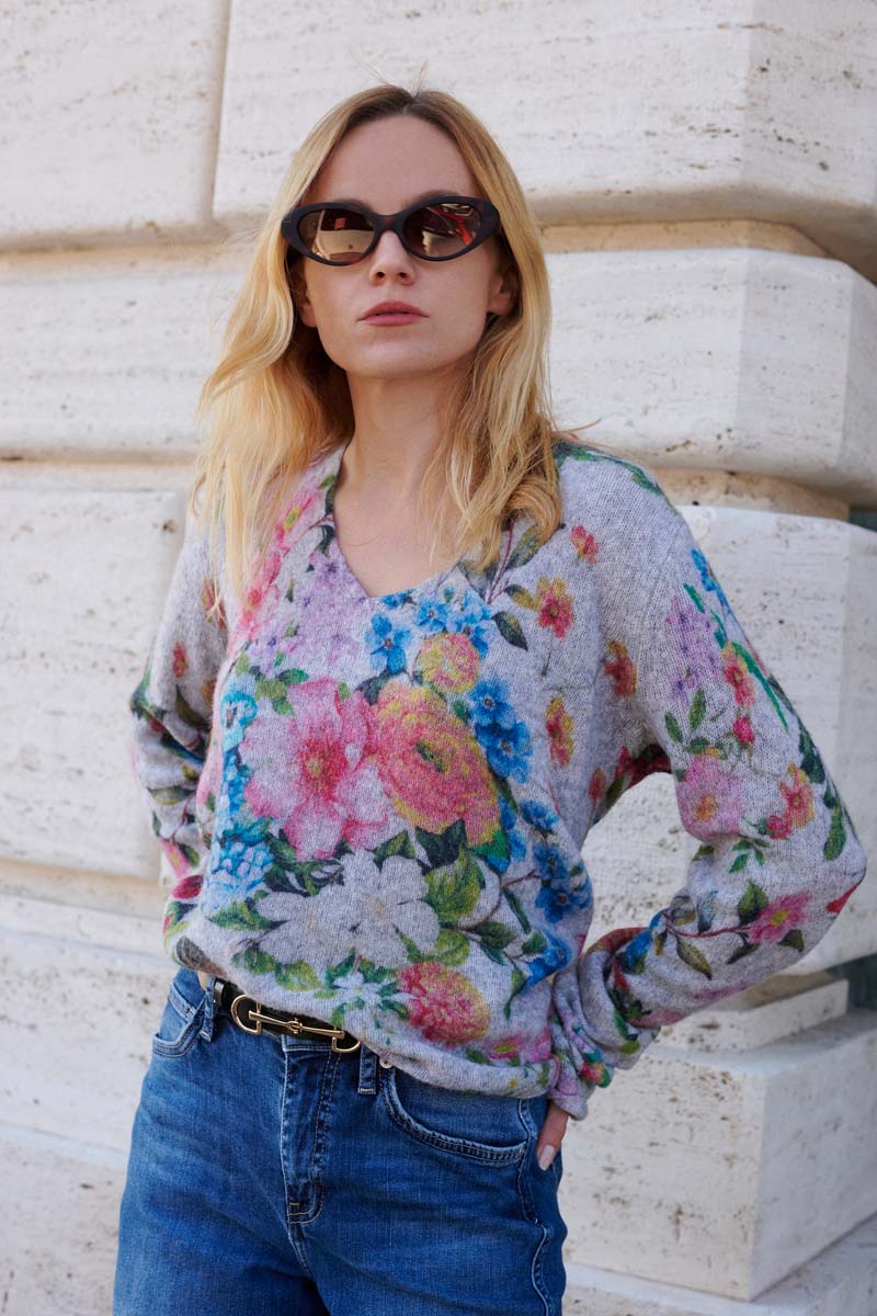 Eine Frau mit langen blonden Haaren trägt eine dunkle Sonnenbrille, einen grauen Princess Goes Hollywood Wollmix-Strickpullover mit buntem Blumendruck und eine blaue Jeans mit schwarzem Gürtel. Sie steht an einer cremefarbenen Steinmauer und blickt mit neutraler Miene leicht nach oben.