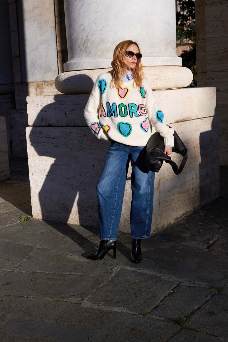 Eine Frau steht im Freien an einer Steinsäule im Sonnenlicht. Sie trägt eine Princess Goes Hollywood-Sonnenbrille, einen weißen Grobstrickpullover aus einem Wollgemisch mit bunten Herzen und dem Schriftzug AMORE" auf der Vorderseite, weite Bluejeans, schwarze Ankle Boots und eine schwarze Tasche.