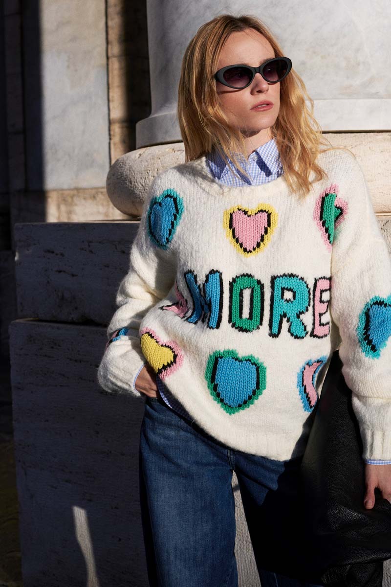 Eine Frau steht draußen an einer Steinmauer. Sie trägt eine dunkle Sonnenbrille und einen handgestrickten weißen Wollmix-Pullover mit bunten Herzen und der Aufschrift "AMORE" über einem blauen Kragenhemd und Jeans. Das Sonnenlicht beleuchtet ihr Gesicht und ihr blondes Haar.