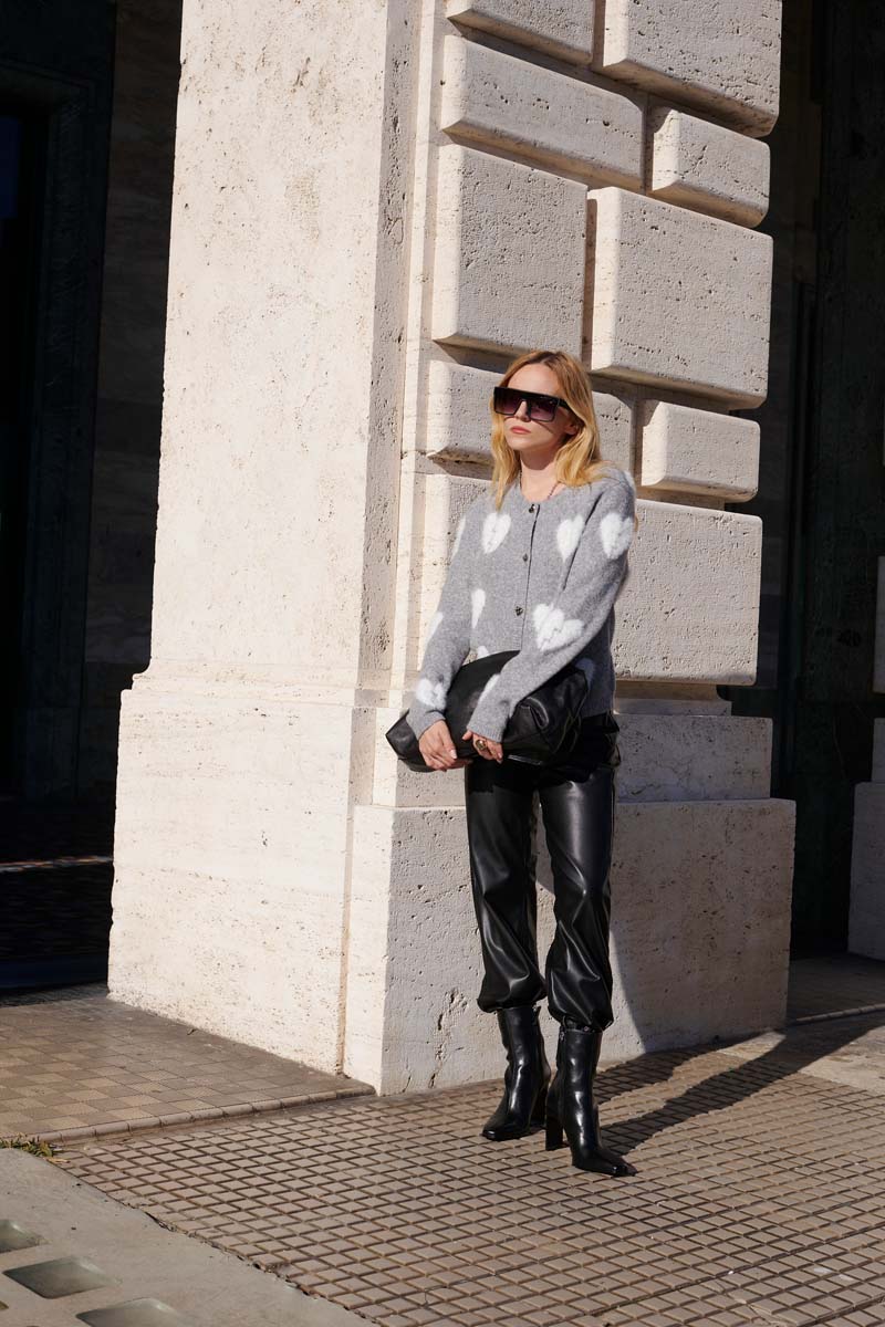 Eine Frau steht draußen an einer Steinmauer. Sie trägt eine graue Wollmix-Strickjacke mit großen weißen Tupfen, eine schwarze Lederhose, hochhackige Stiefel, eine Sonnenbrille und hält eine schwarze Clutch in der prallen Sonne.