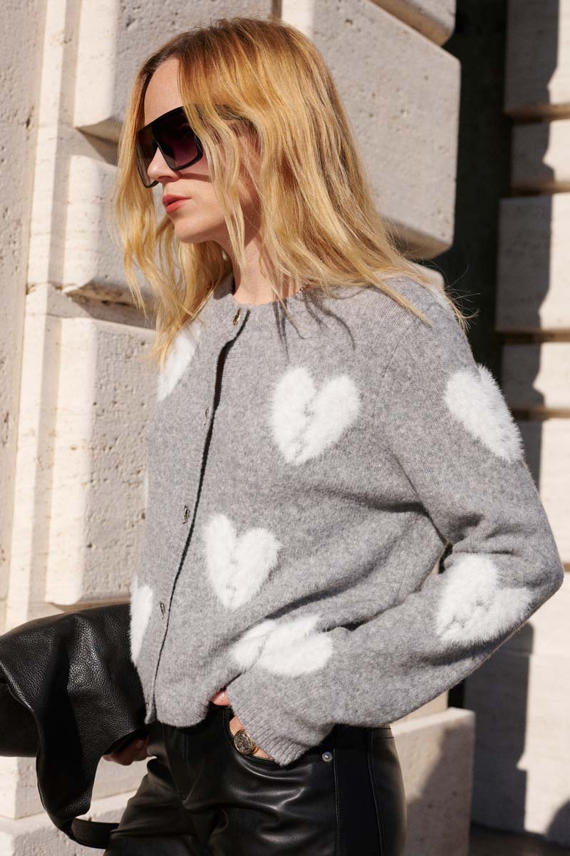 Eine Frau mit langen blonden Haaren und großer dunkler Sonnenbrille trägt eine Princess Goes Hollywood graue Strickjacke aus Wollmix mit weißen Herz-Motiven, steht an einer Steinmauer, hält eine schwarze Lederjacke und trägt eine schwarze Lederhose im Sonnenlicht.